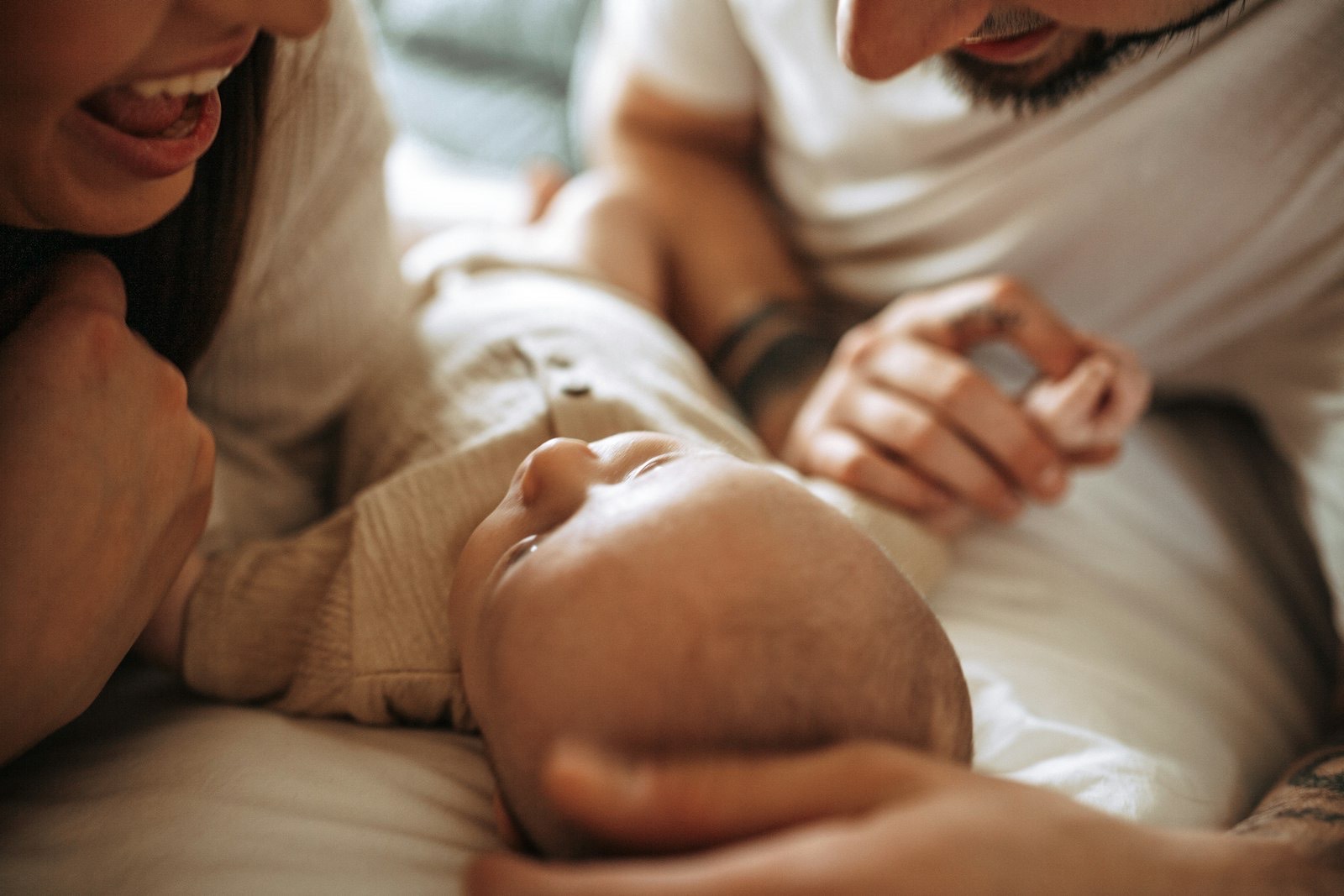 Parents lean over their newborn, laughing softly together on a bed.