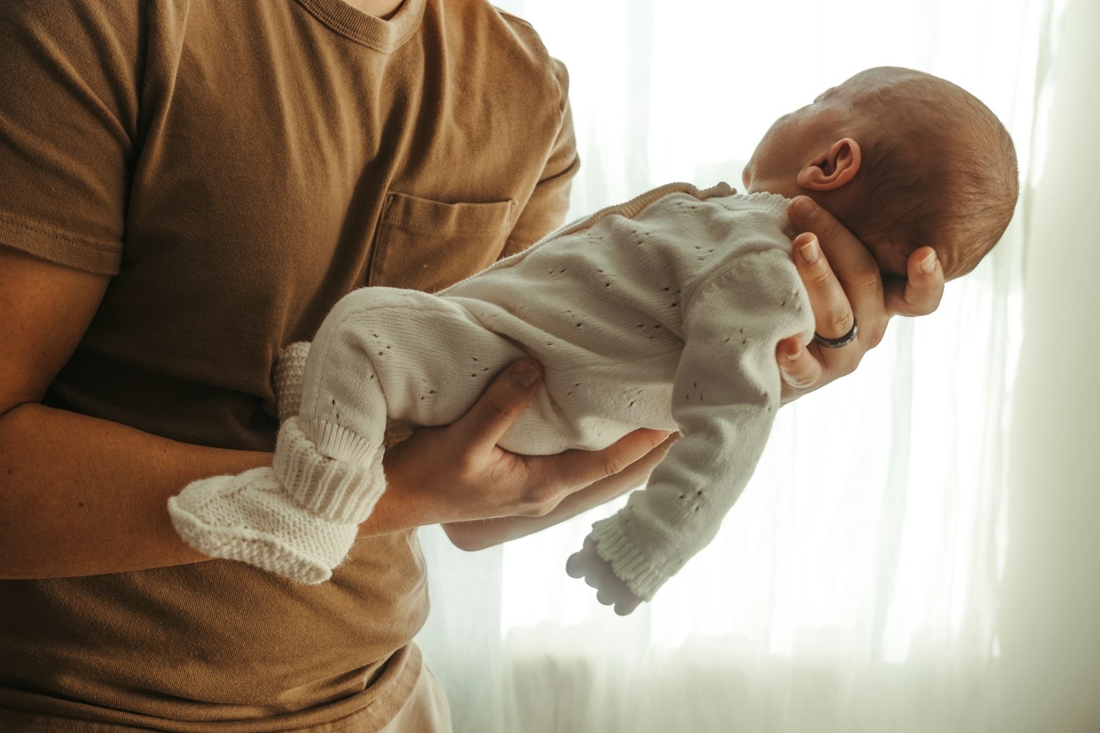 A father gently holds his newborn in a knit onesie by a bright window.