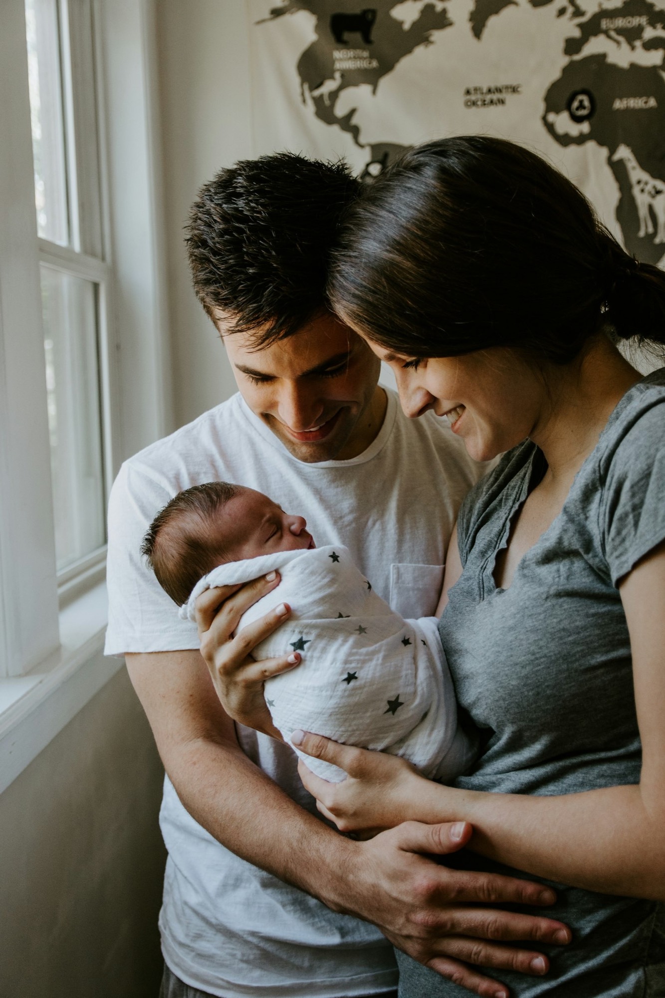 A mother and father smiling down at their swaddled newborn by a bright window.