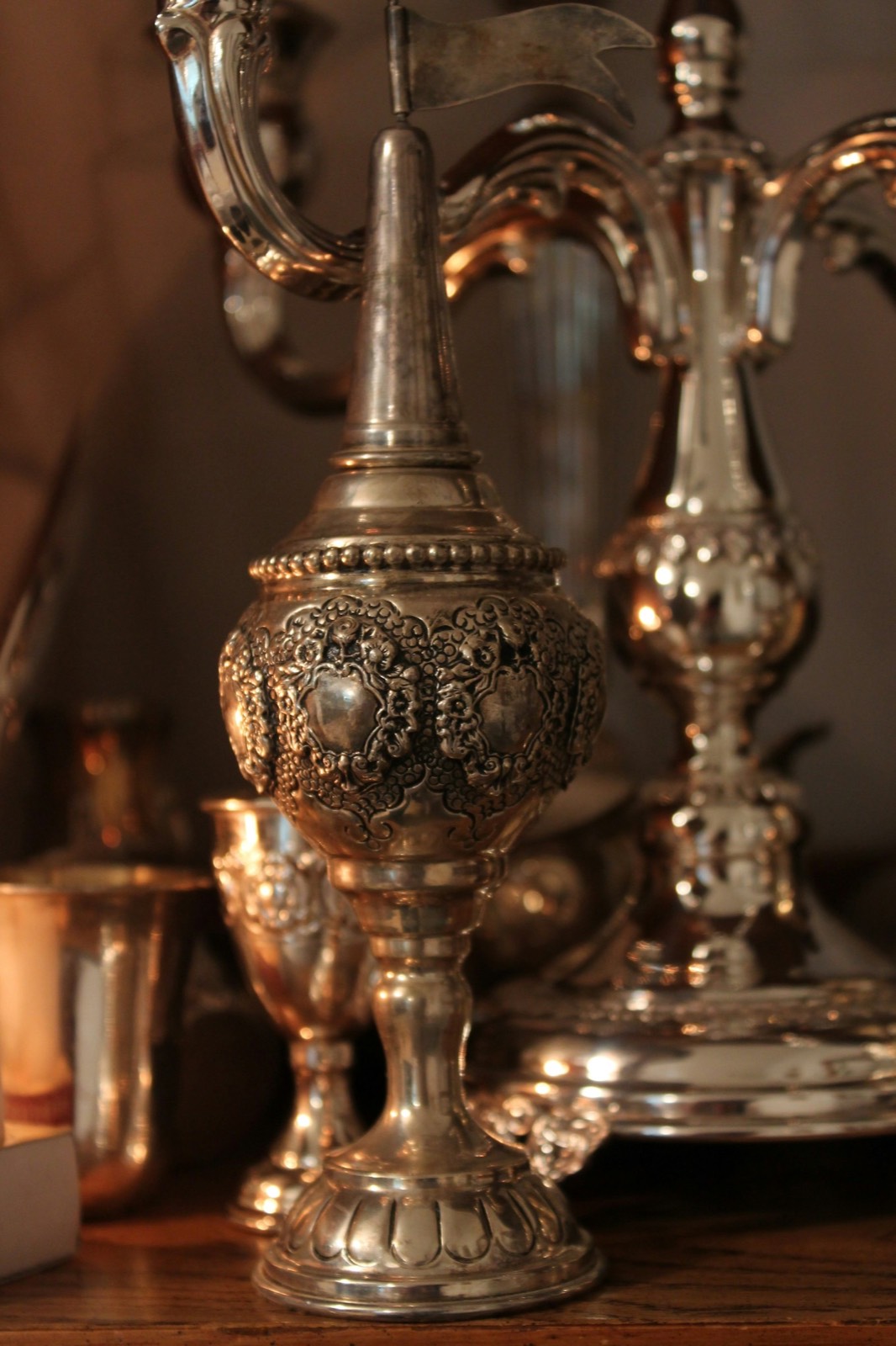 Antique silver Judaica — a besamim spice tower, kiddush cups, and candelabra in warm light.